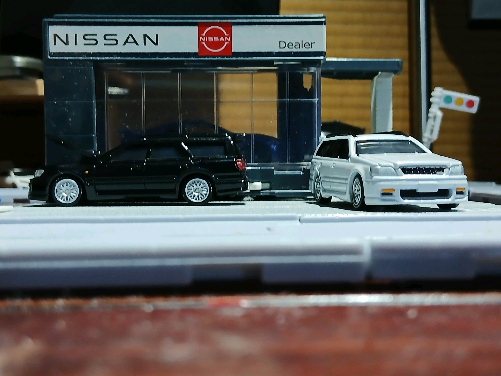 日産ディーラーのミニチュア展示場の前に、発売された日産ステージア 2.5RSの黒と白のミニカー2台が並ぶ様子。家族向けスポーツワゴンを紹介する文脈に合った構図。NISSAN、NISSAN、Dealer