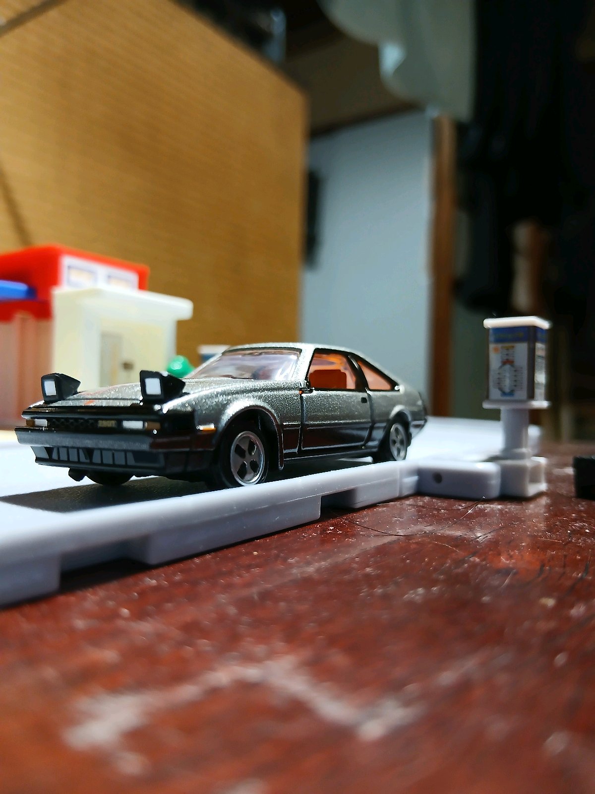 投稿が遅れたという文脈のセリカXXのミニカーで、銀色ボディにリトラクタブルライトを開けた状態の車体が、室内の卓上に置かれた白い道路パーツと小さな標識の横に展示されている