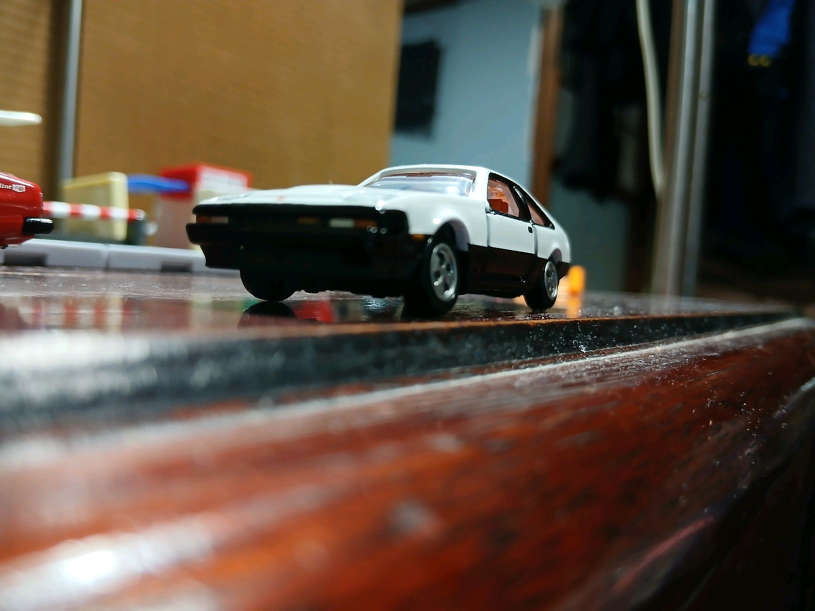 木製テーブルの上に置かれた白と黒のセリカXXのミニカーを、低い位置から手前大きく撮影した写真。背景には赤いミニカーや小物がぼけて写り、投稿文脈どおり遅れて紹介された車両の雰囲気が伝わる。