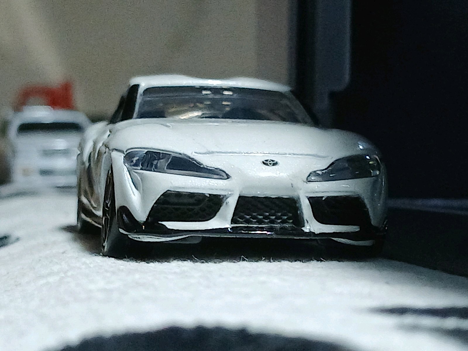 白いトヨタGRスープラのミニカーを正面から捉えた接写で、左後方に別の白い車もぼんやり写る。投稿文脈ではJZA90ではなくBMW由来のDB90として紹介されている。